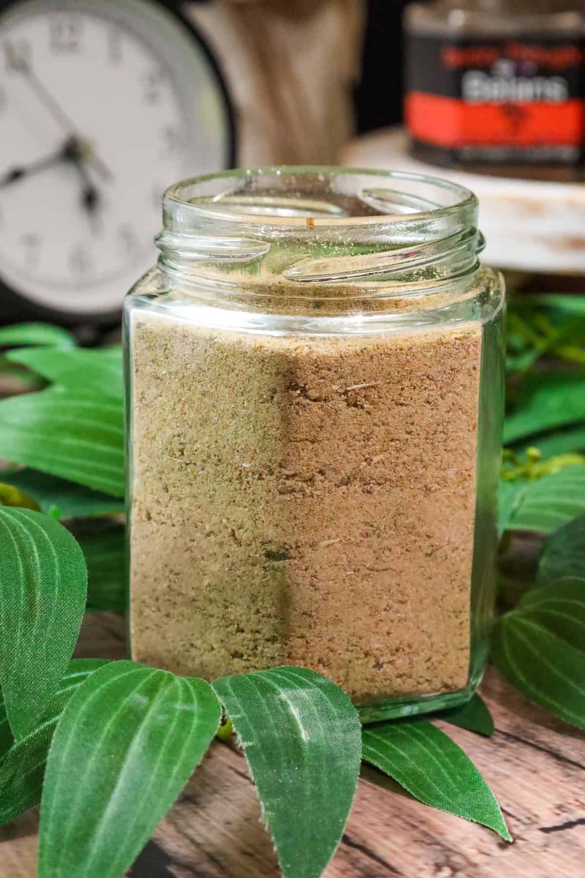 Delmas seasoning in glass jar wihtout a lid on top of greenery with a clock and another seasoning jar in the background.