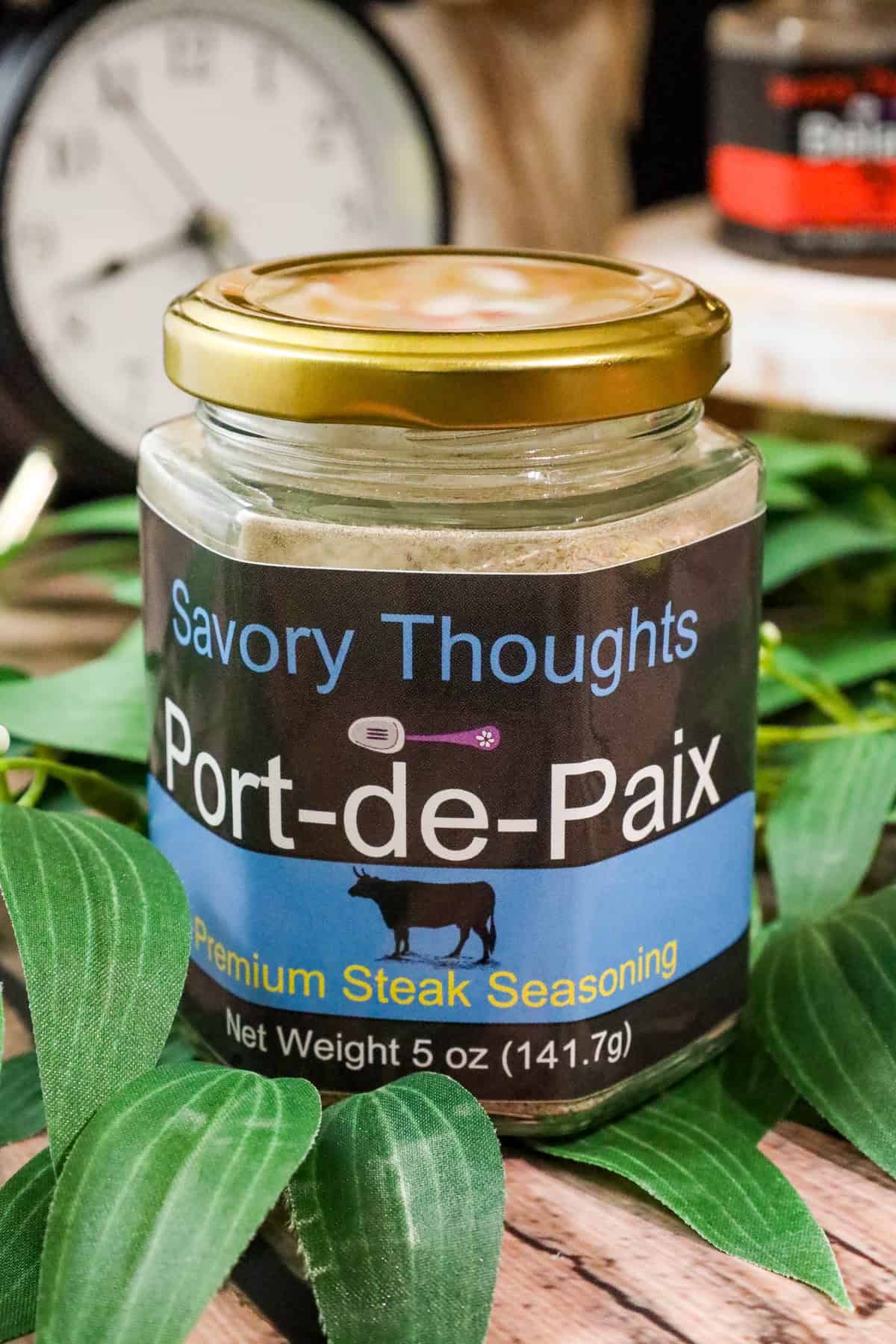 Port-de-Paix seasoning in glass jar on top of wooden table with greenery and clock in background with other barely visible spices.