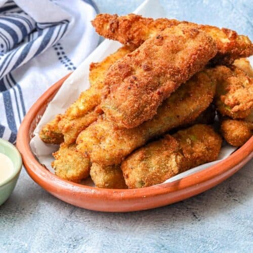 fried pickle spears layered on top of each other in a brown plate with dipping sauce in the background