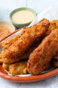 fried pickle spears layered on top of each other on a brown plate with dipping sauce in the background