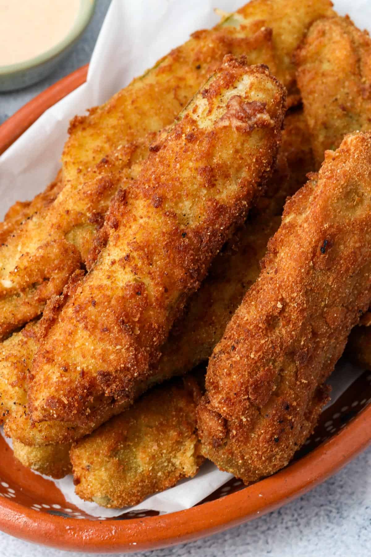 fried pickle spears layered on top of each other on a brown plate with dipping sauce in the background