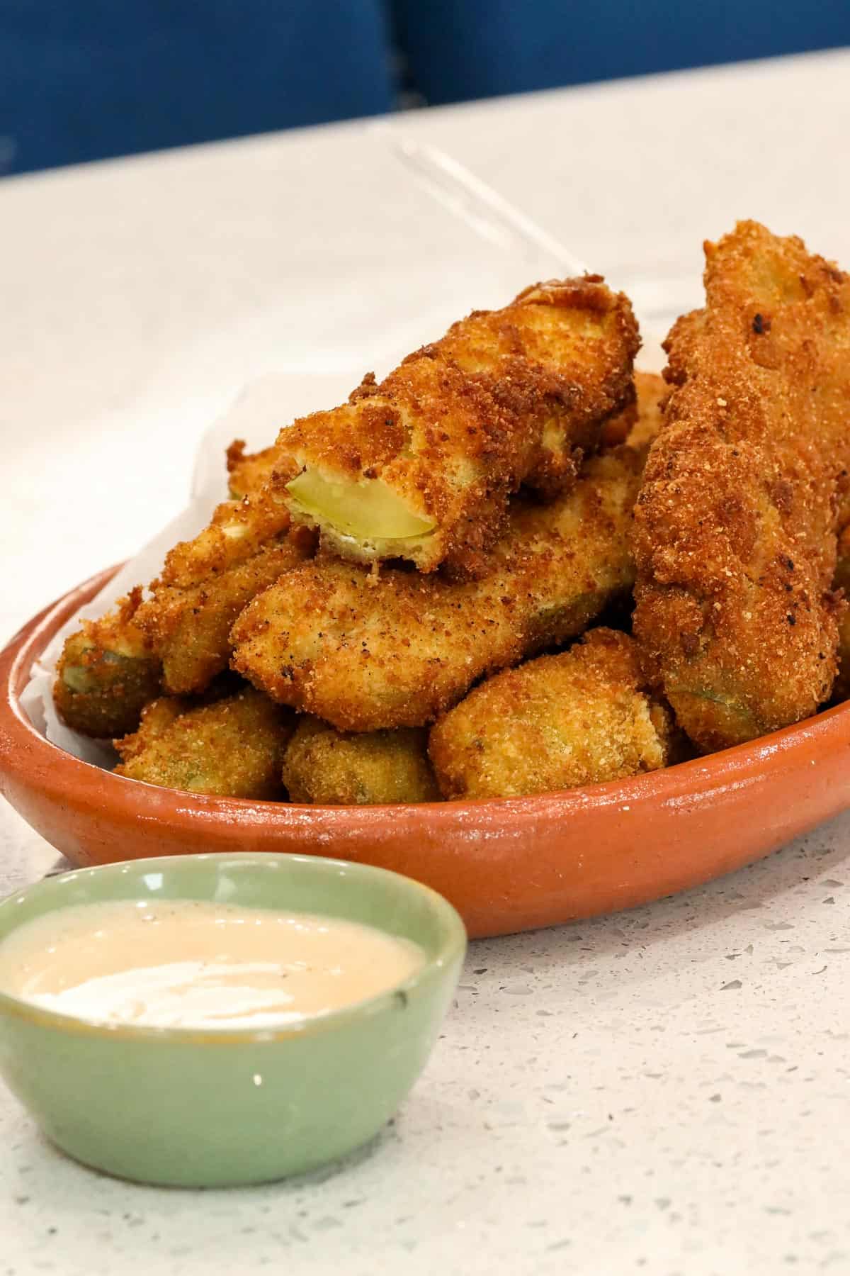 deep fried pickle spears in brown plate on counter with sauce on the side in a green small bowl