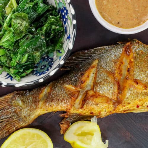 Air Fryer branzino fish on wooden board with dipping sauce, lemon wedges, and sautéed greens