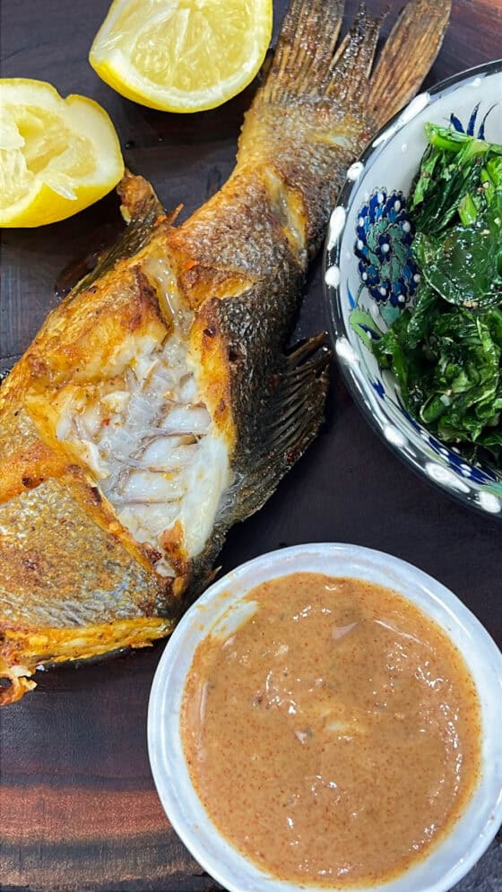 air fryer branzino on wooden board with dipping sauce, greens, and lemon wedges with a bite piece missing from the fish