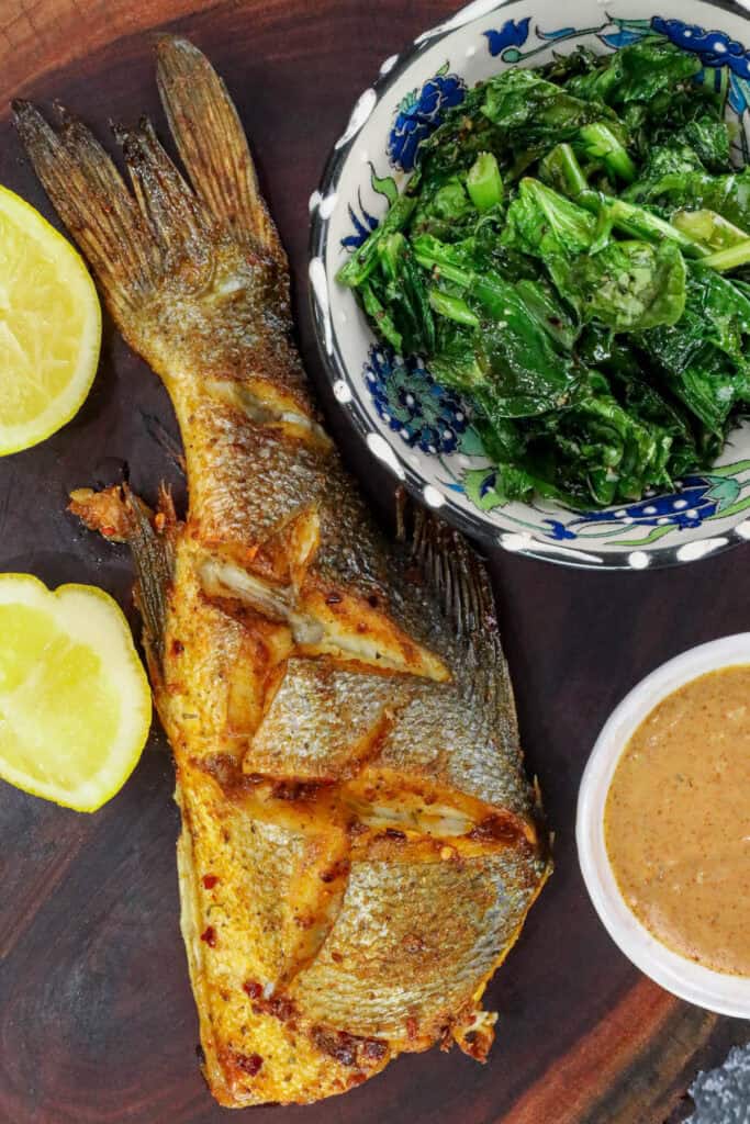 Air Fryer branzino fish on wooden board with dipping sauce, lemon wedges, and sautéed greens